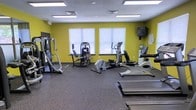 Located at 3201 Walnut Creek Pkwy in Raleigh, NC, this fitness center offers a well-equipped, single-floor workout facility designed to support a range of exercise routines. The space features bold yellow accent walls that create an energizing atmosphere, complemented by bright fluorescent lighting and rubberized flooring that provides both durability and comfort underfoot. Natural light filters in through multiple windows fitted with blinds, adding to the open and inviting feel of the room. The equipment layout is thoughtfully arranged to maximize usable floor space, offering residents easy access to a selection of cardio machines including treadmills, ellipticals, and a stationary bike, as well as strength-training equipment such as a cable machine and a leg press/extension unit. A wall-mounted television is also present, allowing users to stay entertained during their workouts. Informational signage is posted on the walls, keeping users informed of facility guidelines. The overall ambiance is clean, functional, and motivating, making this fitness center a practical and accessible amenity for those looking to maintain an active lifestyle.