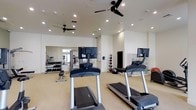 This fitness center offers a comprehensive workout environment designed to accommodate a wide range of exercise needs. The space is finished with clean white walls and a light-toned floor, creating a bright and energizing atmosphere. Recessed lighting throughout the ceiling ensures consistent illumination across the entire area, while multiple ceiling fans promote air circulation and comfort during workouts.

The cardio zone is well-appointed with multiple treadmills, each equipped with individual mounted monitors for entertainment or workout tracking. A large floor-to-ceiling mirror spans one wall, enhancing the sense of space and allowing members to monitor their form during exercise.

The free weights section features a full dumbbell rack with an organized selection of weights, accompanied by a workout bench for strength training exercises. A dedicated functional training area includes a variety of medicine balls and stability balls neatly stored along the wall, catering to dynamic and core-focused workouts. Modular shelving units provide additional organized storage for equipment.

Overall, the fitness center presents a clean, modern, and well-organized environment suited for both cardio and strength training, making it a versatile and fully functional amenity space.