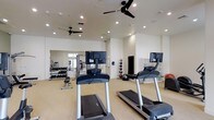 This fitness center offers a comprehensive workout environment designed to accommodate a wide range of exercise needs. The space is finished with clean white walls and a light-toned floor, creating a bright and energizing atmosphere. Recessed lighting throughout the ceiling ensures consistent illumination across the entire area, while multiple ceiling fans promote air circulation and comfort during workouts.

The cardio zone is well-appointed with multiple treadmills, each equipped with individual mounted monitors for entertainment or workout tracking. A large floor-to-ceiling mirror spans one wall, enhancing the sense of space and allowing members to monitor their form during exercise.

The free weights section features a full dumbbell rack with an organized selection of weights, accompanied by a workout bench for strength training exercises. A dedicated functional training area includes a variety of medicine balls and stability balls neatly stored along the wall, catering to dynamic and core-focused workouts. Modular shelving units provide additional organized storage for equipment.

Overall, the fitness center presents a clean, modern, and well-organized environment suited for both cardio and strength training, making it a versatile and fully functional amenity space.