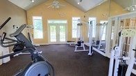 This fitness center offers a spacious and inviting environment designed to support a well-rounded workout routine. The room features a striking vaulted ceiling with a ceiling fan overhead, enhancing air circulation and contributing to a sense of openness throughout the space. Warm yellow walls give the room an energetic and welcoming atmosphere, while durable rubber flooring provides a practical, cushioned surface suitable for a range of exercises.

Natural light flows generously into the space through multiple large windows and a set of glass-paneled double doors that open to an outdoor area, keeping the environment bright and airy. The windows are positioned at varying heights along the walls, including transom-style windows near the roofline, further amplifying the sense of volume and light.

The fitness center is equipped with a cable resistance machine and additional strength training equipment, along with cardio machines such as an elliptical. Wall-mounted mirrors on one side of the room expand the visual depth of the space while allowing users to monitor their form during workouts. Emergency exit signage is clearly posted, reflecting attention to safety throughout the facility.