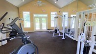 This fitness center offers a spacious and inviting environment designed to support a well-rounded workout routine. The room features a striking vaulted ceiling with a ceiling fan overhead, enhancing air circulation and contributing to a sense of openness throughout the space. Warm yellow walls give the room an energetic and welcoming atmosphere, while durable rubber flooring provides a practical, cushioned surface suitable for a range of exercises.

Natural light flows generously into the space through multiple large windows and a set of glass-paneled double doors that open to an outdoor area, keeping the environment bright and airy. The windows are positioned at varying heights along the walls, including transom-style windows near the roofline, further amplifying the sense of volume and light.

The fitness center is equipped with a cable resistance machine and additional strength training equipment, along with cardio machines such as an elliptical. Wall-mounted mirrors on one side of the room expand the visual depth of the space while allowing users to monitor their form during workouts. Emergency exit signage is clearly posted, reflecting attention to safety throughout the facility.