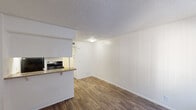 This single-floor unit offers a straightforward, functional layout that makes efficient use of its space. The main living area is bright and open, featuring warm wood-look flooring that runs continuously throughout the unit, crisp white walls, and a textured ceiling. A large window fitted with vertical blinds allows natural light to fill the room, while a nearby entry door provides direct access to the space.

The kitchen is positioned adjacent to the living area and is separated by a half-wall counter with a breakfast bar overhang, creating an open pass-through feel. The galley-style kitchen is equipped with white cabinetry, laminate countertops, a double stainless steel sink, and a black gas range with overhead exhaust hood, offering all the essentials for everyday cooking and meal preparation.

The bathroom continues the wood-look flooring theme and features a wide vanity with a single sink, laminate countertop, ample cabinet storage below, and a large mirror illuminated by a row of vanity lights above. A sliding door leads to an enclosed shower and closet area, maximizing the use of available space.

Overall, this unit presents a clean, move-in ready environment with consistent finishes throughout, offering a practical and comfortable living space.