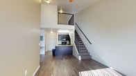 This two-story loft-style residence showcases an open-concept layout that maximizes vertical space with dramatically high ceilings on the main floor. The living area is anchored by warm, wood-look hardwood flooring that runs throughout, creating a cohesive and modern aesthetic. A carpeted staircase with sleek black metal railings leads to the upper-level loft, which overlooks the main floor through a matching black metal balcony railing — adding an industrial-modern design touch to the overall space.

The kitchen is efficiently designed and positioned toward the back of the main floor. It features dark cabinetry, black appliances including a built-in microwave and range, and a peninsula-style breakfast bar with a contrasting dark base that provides additional prep and seating space. Pendant lighting above the kitchen area adds a stylish, contemporary detail.

The upper loft level offers a versatile open space, visible from below and illuminated by a ceiling fan, making it suitable for use as a bedroom, home office, or lounge area. The neutral color palette throughout — warm gray walls paired with light ceilings — creates a bright and airy ambiance. The overall design balances modern industrial elements with practical, comfortable living features across both levels.