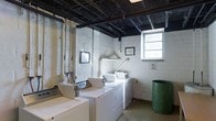 This single-floor space functions as a dedicated laundry room, characterized by its straightforward, utilitarian design. The room features painted white cinder block walls that give it an industrial aesthetic, complemented by an exposed ceiling with visible structural elements, dark-painted beams, and overhead piping running throughout. Multiple top-loading washing machines and dryers are arranged along the walls, providing ample laundry capacity. A small window on the far wall allows natural light to filter into the space, offering ventilation and brightness. The concrete floor and open ceiling infrastructure reinforce the room's functional character. A large green waste bin is positioned in the corner, adding to the practical setup. Ventilation ductwork is visible along the walls, ensuring proper airflow for the appliances. Framed notices or informational signage are mounted on the walls, suggesting shared-use guidelines for occupants. The overall ambiance is straightforward and purposeful, designed entirely around the efficient completion of laundry tasks rather than aesthetic appeal.