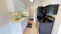 This single-floor 2-bedroom, 1-bathroom residence offers a functional and comfortable layout with a clean, neutral aesthetic throughout. The galley-style kitchen is outfitted with crisp white cabinetry, light countertops, a double-basin sink, and a skylight that brings in natural overhead light. Black appliances, including a range and refrigerator, provide a bold contrast against the white finishes, while light hardwood-style flooring completes the space. An adjacent dining area with a chandelier creates a natural transition between the kitchen and living spaces. The spacious living room is carpeted and filled with natural light from large windows and a sliding glass door that opens to an outdoor area, offering pleasant views of surrounding greenery. A carpeted hallway connects the private quarters, leading to two well-sized bedrooms, each featuring soft carpet, baseboard heating, and windows overlooking lush trees. One bedroom includes a ceiling fan for added comfort. The full bathroom features a tiled tub-shower combination, a granite-topped vanity, and warm overhead lighting. A carpeted hallway with clean white walls ties all rooms together, providing a cohesive and move-in-ready living environment.