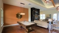 This single-floor space combines a recreation area and living room into one open, versatile layout. A centerpiece pool table with a dark wood pedestal base sits beneath a rectangular pendant light, anchoring the entertainment zone. An accent wall painted in a warm terracotta tone adds visual depth and contrast to the otherwise neutral color palette. Adjacent to the game area, a built-in dark wood shelving unit frames a stone fireplace, creating a striking focal point. The fireplace surround features stacked stonework that adds a rustic, textured element to the space. A flat-screen television is mounted within the shelving unit, seamlessly integrating media into the design. Polished concrete flooring in a rich, dark hue runs throughout, lending an industrial yet refined character to the room. A sunburst mirror decorates the accent wall, adding an artistic touch. The living area includes a neutral-toned sofa positioned near a window that allows natural light to filter in. The overall ambiance is warm, inviting, and well-suited for entertaining.