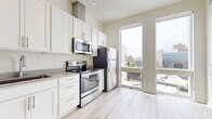 This single-floor unit showcases a thoughtfully designed interior with a contemporary aesthetic, highlighted by light hardwood flooring and a neutral color palette that creates a bright, airy atmosphere. The kitchen is a standout feature, offering an abundance of white shaker-style cabinetry that extends to the ceiling, providing generous storage throughout. A full suite of stainless steel appliances includes a French door refrigerator, electric range, built-in microwave, and dishwasher, all complemented by sleek countertops and a single-basin sink. Track lighting above adds functional illumination to the workspace. Floor-to-ceiling windows in the living area flood the space with natural light while framing sweeping urban views of the surrounding cityscape, adding a dynamic visual element to the interior. The tall ceilings enhance the sense of openness, contributing to an expansive feel. The bathroom features a clean, minimalist design with a wide white vanity, under-mount sink, built-in storage shelving, and a large mirror, maintaining the unit's consistent modern finish. Overall, the space presents a polished, move-in-ready environment suited for urban living.
