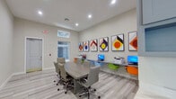 This single-floor business center is thoughtfully designed to support both collaborative work and individual productivity. The main area centers around a spacious rectangular conference table surrounded by ergonomic upholstered chairs, ideal for meetings or group work sessions. Adjacent to the conference area, a built-in wall-mounted workstation runs along one side of the room, equipped with multiple desktop computers, a printer, and colorful modern shell chairs in orange, green, and blue — adding a lively contrast to the otherwise neutral palette.

The interior is finished with light wood-look flooring and warm beige walls, giving the space a clean, professional atmosphere. Recessed ceiling lighting provides consistent, even illumination throughout. A row of bold, framed geometric artworks lines the main wall, featuring vivid shapes in red, orange, blue, and yellow tones that energize the room's aesthetic.

A built-in cabinetry unit with a marble-style countertop offers additional storage and surface space near a glass-paneled door, which leads to an adjacent area filled with natural light. High clerestory windows further enhance the brightness of the space. The layout is open and functional, making this business center well-suited for a variety of professional and collaborative needs.