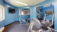 Located at 90 Gerrish Ave in East Haven, Connecticut, this dedicated fitness center offers a thoughtfully arranged exercise space designed to support a full range of workout activities. The facility comprises a main exercise room and a connecting hallway, all situated on a single level.

The exercise room is defined by bold blue walls that create an energizing atmosphere, complemented by durable rubber flooring suited for high-traffic fitness use. Fluorescent overhead lighting ensures the space is well-lit throughout. Multiple windows along one wall invite natural light and offer pleasant views of the surrounding greenery, enhancing the workout environment.

The room is comprehensively equipped with a varied selection of fitness machines and equipment. Cardio options include a treadmill, elliptical trainer, recumbent bike, and upright stationary bike. A multi-station cable and weight machine anchors the strength-training area, which is further supported by a full dumbbell rack with a flat bench. A wall-mounted flat-screen television adds convenience for entertainment during workouts. A wall-mounted air conditioning unit ensures year-round climate comfort. The hallway provides a clean transition into the exercise room, keeping the layout organized and accessible. Overall, the space delivers a well-rounded, functional fitness facility.