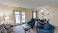 This single-floor residence offers a comfortable and well-appointed living environment with a thoughtful layout designed for everyday ease. The combined living and dining area serves as the heart of the home, bathed in natural light streaming through large French doors that open to a lush outdoor view, as well as an additional side window dressed with elegant curtain panels. The living area features plush wall-to-wall carpeting underfoot and is furnished with classic blue accent chairs accented with nailhead trim, centered around a round glass-top coffee table — creating a relaxed yet refined seating arrangement. A ceiling fan with an integrated light fixture provides both comfort and ambient lighting to the space. Adjacent to the living area, the dining space accommodates a round wooden table with upholstered chairs, ideal for intimate meals and gatherings. The walls are finished in a soft, warm neutral tone that complements the natural light and contributes to the overall airy feel of the space. Framed artwork adds a subtle decorative touch. The seamless flow between the living and dining zones makes this a versatile and welcoming space suited for both relaxing and entertaining.