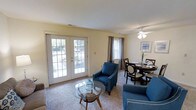 This single-floor residence offers a comfortable and well-appointed living environment with a thoughtful layout designed for everyday ease. The combined living and dining area serves as the heart of the home, bathed in natural light streaming through large French doors that open to a lush outdoor view, as well as an additional side window dressed with elegant curtain panels. The living area features plush wall-to-wall carpeting underfoot and is furnished with classic blue accent chairs accented with nailhead trim, centered around a round glass-top coffee table — creating a relaxed yet refined seating arrangement. A ceiling fan with an integrated light fixture provides both comfort and ambient lighting to the space. Adjacent to the living area, the dining space accommodates a round wooden table with upholstered chairs, ideal for intimate meals and gatherings. The walls are finished in a soft, warm neutral tone that complements the natural light and contributes to the overall airy feel of the space. Framed artwork adds a subtle decorative touch. The seamless flow between the living and dining zones makes this a versatile and welcoming space suited for both relaxing and entertaining.