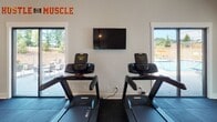 This fitness center is a thoughtfully designed workout facility that combines functionality with an energizing atmosphere. The space is anchored by striking vaulted ceilings accented with exposed dark wood beams, recessed lighting, and ceiling fans that enhance airflow and comfort during workouts. Dark rubber flooring spans the entire area, providing durability and safety underfoot.

The strength training zone is outfitted with a suite of Precor cable and resistance machines, a functional trainer, and a heavy punching bag suspended from the ceiling frame — catering to a wide range of strength and conditioning exercises. The cardio section features multiple treadmills and an elliptical trainer, positioned near large windows that overlook an outdoor pool and patio area, offering natural light and pleasant views while exercising. A wall-mounted flat-screen television is conveniently placed in the cardio zone for entertainment.

Motivational wall graphics and quotes in bold orange and white lettering — including phrases like "Hustle for Muscle" and "Harder. Better. Faster. Stronger." — energize the space and reinforce a focused fitness mindset. Multiple windows and glass doors throughout the facility allow natural light to flood the interior, creating an open and inviting environment. The overall design balances a modern aesthetic with practical, high-quality fitness amenities.