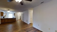 This single-floor space offers a clean, functional layout with a cohesive design throughout. The main living area is grounded by rich, dark hardwood flooring that extends seamlessly through the entire unit, lending warmth and character to the interior. A ceiling fan with an integrated light fixture provides both comfort and illumination in the main living space.

The open layout allows for a natural flow between the living area and the kitchen, which features warm wood cabinetry and a sink along the far wall. The kitchen is compact yet efficiently designed, making use of every inch of available space.

A central hallway connects the main living area to the rest of the unit, lined with white walls and neutral-toned doors that contribute to the bright, airy feel. Along the hallway, practical utility features are visible, including what appears to be a water heater, floor vents for climate control, and a thermostat — all speaking to the unit's functional infrastructure.

The interior is finished with white walls and white trim throughout, providing a neutral backdrop that suits a variety of interior styles. The textured ceiling adds a subtle design element overhead. Overall, the space presents a straightforward and practical layout with durable finishes, making it well-suited for everyday living.