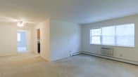 This single-floor space presents a straightforward and functional layout designed for comfortable everyday living. The main living area is generously proportioned, finished with neutral wall-to-wall carpeting and crisp white walls that create a clean, airy atmosphere. A large double-hung window along the exterior wall allows abundant natural light to fill the room while offering views of surrounding greenery, contributing to a bright and pleasant ambiance.

Climate comfort is addressed through a combination of a baseboard heating unit running along the base of the exterior wall and a through-the-wall air conditioning unit positioned beneath the window, ensuring year-round temperature control. A standard electrical outlet is conveniently placed along the baseboard.

A partial wall divides the living area from a connected hallway that leads to the kitchen, which is visible in the background and features wood-toned cabinetry and a dark appliance. The layout encourages an open sightline between spaces while maintaining a degree of separation. The overall aesthetic is simple and neutral, providing a versatile foundation well-suited to a variety of furnishing styles and personal touches.