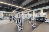 This expansive, single-floor fitness center offers a thoughtfully designed workout environment that balances both function and style. The space is divided into distinct training zones, providing ample room for a comprehensive range of exercise activities without feeling crowded.

The architectural design features a striking exposed ceiling with dark-painted ductwork and structural elements, contrasted by bold linear LED lighting that runs across the ceiling in geometric patterns, creating a dynamic and energizing atmosphere. Large floor-to-ceiling glass garage-style doors and windows line the perimeter, flooding the space with abundant natural light and offering open views of the surrounding outdoor greenery.

The strength training area is well-appointed with a full array of selectorized cable machines, a complete dumbbell rack ranging across multiple weight increments, adjustable benches, and functional training rigs. Large mirrors along the walls enhance the sense of space and allow for proper form checks during workouts.

The cardio zone features multiple treadmills, including curved manual models and motorized units, as well as stair climbers, all positioned to take advantage of the natural light and outdoor views. A dedicated functional training corner stocks medicine balls, kettlebells, resistance bands, and a wall-mounted training rig. Glass partition walls elegantly divide sections of the space while maintaining visual openness throughout. Durable rubber flooring completes this professional-grade facility.