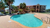 This single-floor unit is part of a Mediterranean-inspired residential community characterized by warm earth-toned exteriors, red-trimmed rooflines, arched entryways, and private balconies. The community's standout outdoor amenity is a generously sized freeform pool with crystal-clear turquoise water, framed by a decorative rock waterfall feature, mature palm trees, and a wide brick-paved sun deck furnished with lounge chairs — creating a resort-like atmosphere ideal for relaxation.

The interior of the unit features warm, neutral-toned walls that contribute to a clean and inviting ambiance throughout. Rich, dark hardwood-style flooring runs consistently across the rooms, providing a grounding contrast to the light-colored walls and white trim. The rooms are well-proportioned, with natural light entering through windows fitted with horizontal blinds, offering both brightness and privacy control. Six-panel white interior doors with brushed nickel hardware connect the living spaces, while white baseboards provide a crisp finishing detail. Closet access is visible in at least one room, adding practical storage. The overall interior presents a straightforward, move-in-ready aesthetic with a cohesive and understated design sensibility.