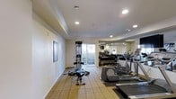 This single-floor fitness room offers a comprehensive workout environment designed to accommodate a variety of exercise routines. The space is outfitted with a selection of cardio equipment, including elliptical trainers and treadmills, positioned along one side of the room for convenient use. A multi-function cable weight station anchors the center of the room, complemented by a free weight rack visible along the back wall, offering users a full range of strength training options.

The room features elegant crown molding along the ceiling perimeter, adding a refined architectural touch that elevates the space beyond a standard gym environment. Recessed lighting throughout provides bright, even illumination, while a large window along the back wall invites natural light and offers a view of the surrounding greenery outdoors. Light-toned tile flooring and neutral walls contribute to an open, clean aesthetic. A floor-to-ceiling mirror along one wall enhances the sense of spaciousness and allows users to monitor their form during workouts. The overall ambiance is functional yet polished.