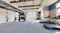 Located at 1421 Rural Hill Rd in Nashville, TN, this generously sized fitness center offers a well-appointed, single-floor workout facility designed to accommodate a wide range of exercise needs. The space makes an immediate impression with its soaring coffered ceiling accented by rustic exposed wood beams, recessed lighting, and dual ceiling fans that ensure a comfortable environment throughout.

Soft blue-gray walls contrast with crisp white trim and wainscoting, lending the space a clean, modern aesthetic. Plush blue-toned carpet covers the entire floor, providing cushioning underfoot. Expansive banks of large windows line multiple walls, flooding the interior with natural light and offering views of the surrounding landscaping and grounds.

The fitness floor is organized to support diverse training styles. A dedicated strength zone features a TRX suspension system, a full power rack loaded with weight plates, a multi-functional cable machine, and a tiered dumbbell rack. Cardio equipment includes multiple Spirit treadmills arranged in a row for group or individual sessions. A functional training area is equipped with plyometric boxes, medicine balls, stability balls, and adjustable benches. Full-length mirrors along one wall enhance the sense of space and allow for form checks during workouts. A wall-mounted flat-screen TV and glass-panel entry doors with adjacent storage complete this well-rounded facility.