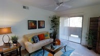 This single-floor residence offers a clean, modern aesthetic with a thoughtfully arranged layout designed for comfortable everyday living. The living room serves as the heart of the space, featuring light-toned walls that create an airy, open atmosphere. A ceiling fan is centrally positioned overhead, providing both functional airflow and a subtle design element. Natural light fills the room through a large sliding glass door fitted with vertical blinds, offering a seamless connection to an outdoor area beyond. The living room is furnished with a plush sofa accented by vibrant decorative pillows in orange, teal, and yellow, complemented by a glass-top coffee table and a colorful area rug that adds a lively touch to the flooring. A tall decorative plant adds a natural, organic element to the corner of the room, while a pair of framed wall art pieces provide visual interest above the sofa. The overall ambiance of the space is bright, casual, and inviting, with a contemporary décor style that balances comfort and visual appeal. The presence of a sliding door leading to an exterior space suggests the residence benefits from indoor-outdoor connectivity, enhancing its livability and sense of openness.