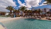 This outdoor amenity space in Jacksonville, FL, is designed with a resort-inspired aesthetic that seamlessly blends indoor comfort with Florida's sunny climate. The centerpiece is a generously sized swimming pool with clear, sparkling water, framed by a wide pool deck lined with rows of chaise lounges ideal for sunbathing. Tall palm trees surround the area, providing a tropical atmosphere beneath open, sun-filled skies.

Adjacent to the pool is a spacious covered pavilion with a warm wood-paneled ceiling and large ceiling fans that maintain airflow throughout. The pavilion features distinct seating zones, including a stylish lounge area anchored by a floor-to-ceiling stone fireplace wall with a wall-mounted TV, creating an inviting social hub. An outdoor dining area accommodates group gatherings, while additional casual seating clusters offer more intimate settings.

Large floor-to-ceiling windows and glass doors along the pavilion's interior-facing wall allow natural light to flow freely, visually connecting the indoor and outdoor areas. Neutral tile flooring, clean white walls, and contemporary woven furniture throughout contribute to a cohesive, modern coastal design. The overall ambiance is relaxed yet refined, offering a welcoming retreat for residents.