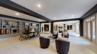Located at 9015 Element Cir in Garner, NC, this single-floor clubhouse facility presents a refined, contemporary design characterized by a bold contrast of dark charcoal accents against crisp white walls and light-toned large-format tile flooring accented with rich dark marble inlays. The expansive main lounge area features coffered and beam-detailed ceilings, recessed lighting, and multiple seating configurations — from plush barrel chairs to a tufted leather sofa — creating versatile gathering spaces. An eye-catching chandelier anchors a more intimate lounge zone, while built-in shelving displays curated décor throughout. The communal kitchen is a standout feature, offering a generous island with bar seating, white shaker cabinetry, a textured mosaic backsplash, stainless steel appliances, and a wall-mounted television. A separate media lounge features bold teal walls adorned with a gallery-style art arrangement and a large sectional sofa, offering a cozy retreat. The entry corridor showcases dramatic tray ceilings, large-scale artwork, and polished flooring that sets an elegant tone. Expansive glass doors and oversized windows flood the space with natural light and provide direct access to an outdoor patio, seamlessly connecting indoor and outdoor living.