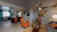 This leasing office is a well-appointed, single-floor space designed to be both functional and inviting. The layout flows naturally between two distinct areas: a comfortable lounge and a professional workspace, connected by an open passageway that gives the office a spacious, airy feel.

The lounge area sets an immediately welcoming tone, furnished with a plush gray sofa and bold orange accent chairs arranged around a glass-topped coffee table. A vivid patterned area rug in warm golds and teals anchors the seating arrangement, while colorful abstract artwork on the walls adds energy and personality. A ceiling fan with integrated lighting adds a practical touch overhead, and natural light filters in through nearby windows.

Adjacent to the lounge, a dedicated office area provides a fully equipped workspace with a large dark desk, ergonomic seating, and ample storage through additional dark-toned cabinetry and a multi-drawer dresser. Teal curtains frame the window, adding a pop of color that ties back to the lounge's palette. Further abstract artwork adorns the walls throughout, maintaining a cohesive and vibrant aesthetic.

Light wood-tone flooring runs throughout the entire space, complementing the neutral gray walls and creating a clean, modern backdrop. A tall indoor fiddle-leaf fig plant and decorative greenery add natural warmth to the environment, making the office feel approachable and lively for prospective residents.