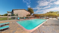 Located at 715 Marie Antoinette St. in Lafayette, Louisiana, this outdoor pool area offers a well-appointed recreational retreat designed for comfort and leisure. The centerpiece is a generously sized rectangular swimming pool with crystal-clear turquoise water, framed by a durable brick paver deck that surrounds the entire perimeter. The pool is enclosed by a wrought iron fence with brick pillar accents, providing both security and an attractive boundary that opens to views of manicured green lawns and mature trees beyond.

Multiple lounge chairs are positioned around the pool deck, offering ample space for sunbathing and relaxation. A dedicated poolside structure with a covered patio and large sliding glass doors serves as a convenient amenity building, providing shade and shelter adjacent to the water. Additional seating areas with chairs and tables are scattered throughout the deck, accommodating both individual lounging and small gatherings.

The surrounding landscape includes flowering trees and established shade trees that add natural color and character to the environment. The brick exterior of the adjacent multi-story building complements the warm tones of the paver deck, creating a cohesive architectural aesthetic. Under open Louisiana skies, this pool area presents a functional and inviting outdoor space well-suited for warm-weather enjoyment.