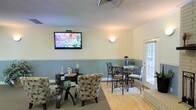 This single-floor space is designed to offer a comfortable and inviting atmosphere for visitors. The main lounge area features a thoughtfully curated mix of seating options, including plush patterned accent chairs arranged around a glass-top table, as well as bistro-style high tables with wrought iron chairs, providing flexibility for both relaxed and upright seating preferences. A wall-mounted flat-screen television serves as a focal point on one wall, ideal for keeping guests informed or entertained. The interior design showcases a two-tone wall treatment, with soft sage green on the lower half and a neutral cream tone above, creating a calming, professional aesthetic. A wall sconce adds a warm, ambient glow to complement the natural light streaming in through a paneled exterior window and door. The flooring transitions from hardwood to a neutral area rug, defining the separate seating zones while maintaining a cohesive look throughout. Informational brochures and decorative floral accents are thoughtfully placed to enhance the welcoming character of the space. The overall ambiance is clean, organized, and serene — well-suited for a professional service environment such as a medical, wellness, or business office.