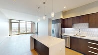 Located in the heart of downtown Kalamazoo at 180 E Water Street, this contemporary single-floor unit offers a thoughtfully designed open-concept layout finished with light hardwood flooring throughout. The kitchen serves as a central focal point, boasting rich dark-toned cabinetry, a geometric-patterned tile backsplash, quartz countertops, stainless steel appliances, and a large center island illuminated by sculptural pendant lighting — ideal for both cooking and entertaining. The living area is generously proportioned, bathed in natural light streaming through floor-to-ceiling windows and sliding glass doors that open to a private balcony, offering elevated views of the surrounding Kalamazoo cityscape. The bedroom features its own balcony access via sliding glass doors, a walk-in closet, and a ceiling fan, creating a comfortable and airy retreat. Two bathrooms complete the layout: one featuring a frameless glass walk-in shower with a built-in niche and stone tile flooring, and a second offering a full soaking tub and modern vanity with integrated lighting. Throughout the space, clean architectural lines, recessed lighting, dark door frames, and a neutral color palette contribute to a cohesive, upscale modern aesthetic.