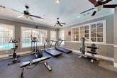 Located at 100 Ardsley Place in Norcross, Georgia, this fitness center offers a fully equipped workout space designed for a comprehensive exercise experience. The room features a warm, neutral color palette with gray walls accented by crisp white crown molding and white chair rail trim, lending the space a polished, upscale feel. Multiple ceiling fans are distributed throughout the room, ensuring consistent airflow and comfort during workouts, complemented by recessed lighting overhead.

The cardio zone is well-stocked with multiple treadmills, elliptical machines, and a rowing machine, all positioned near a bank of large windows that frame views of the outdoor pool area, providing natural light and an open, airy atmosphere. The strength and resistance training area includes a multi-function cable machine, a dumbbell rack, weight benches, and a leg extension/curl machine, all arranged on protective rubber flooring mats set over durable carpet. A large wall mirror expands the visual depth of the space and helps users monitor their form during exercises.

The fitness center is accessible via a dedicated entry door and features interior French-style windows that connect visually to adjacent common areas. Safety signage and an emergency exit are clearly marked. Overall, this fitness center delivers a functional, comfortable, and well-appointed environment suited to a variety of workout routines.