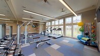 This fitness center is a thoughtfully designed, single-floor space that offers a comprehensive workout environment. The room is defined by its tall, floor-to-ceiling windows along one wall, which flood the interior with natural light and provide open views of the surrounding outdoor greenery and neighboring buildings. The patterned carpet flooring, composed of blue, gray, and beige modular tiles arranged in a block design, adds a contemporary aesthetic while providing cushioned support underfoot.

The ceiling features recessed linear LED lighting strips and multiple ceiling fans, ensuring even illumination and comfortable air circulation throughout the space. Full-length mirrors are mounted along one wall, visually expanding the room and aiding exercisers in monitoring their form.

The cardio section is well-stocked with multiple treadmills and elliptical trainers positioned near the windows, taking advantage of the natural light and outdoor views. The strength training area includes a cable machine, weight bench, and a rack of dumbbells in graduated sizes. A built-in open shelving unit along the back wall provides organized storage, and a wall-mounted television is available for entertainment. Additional accessories, including stability balls, medicine balls, resistance bands, and foam rollers, are neatly stored on a dedicated rack, rounding out the fitness offerings of this space.