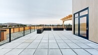 Located at 2826 Jacklin Rd in Langford, Victoria, BC, this rooftop terrace is a thoughtfully designed outdoor amenity space offering sweeping panoramic views of the surrounding mountains, treeline, and distant waterway. The terrace is finished with large-format concrete pavers laid in a clean grid pattern, providing a durable and visually cohesive surface across the expansive open-air level.

A standout architectural feature is the natural wood pergola, constructed from warm-toned timber, which serves as a shaded gathering focal point and adds an organic contrast to the modern building exterior. The exterior cladding of the rooftop stairwell enclosure features light wood-toned paneling, complementing the pergola's aesthetic and contributing to the overall contemporary design language.

Several large, dark metal raised planter boxes are positioned across the terrace, offering opportunities for greenery and landscaping. Black steel railings with glass and vertical bar infill panels define the perimeter, ensuring unobstructed sightlines to the surrounding landscape while maintaining safety. Exterior wall-mounted lighting fixtures provide illumination for evening use. Access to the terrace is provided via a clearly marked stairwell entry door. The space is well-suited for community gathering, outdoor events, or relaxed enjoyment of Victoria's natural scenery.