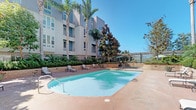 This thoughtfully designed outdoor space offers a seamless blend of resort-style amenities and lush, manicured landscaping. At the heart of the space is a generously sized swimming pool with crystal-clear water, surrounded by a spacious pool deck finished in warm-toned pavers. Sun loungers are arranged along the poolside, providing ample space for relaxation beneath the open sky. Tall palm trees and mature magnolias frame the building's modern facade, enhancing the sense of a private retreat.

Adjacent to the pool, a beautifully paved courtyard serves as an inviting outdoor lounge. The stone-tiled patio features wicker furniture with striped cushions arranged in conversational groupings, creating a comfortable and welcoming atmosphere. A curved raised planter border, finished in textured masonry, encloses a rich variety of tropical plants, ferns, and flowering shrubs that lend vibrant color and a natural sense of enclosure to the space.

Large glass doors and windows along the building's ground-floor facade blur the boundary between interior and exterior, flooding adjacent indoor spaces with natural light. The overall ambiance is serene and resort-inspired, offering a private garden-like escape enhanced by mature trees, layered greenery, and carefully considered outdoor furnishings.