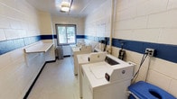This shared laundry facility is a practical, well-equipped space designed to serve multiple residents. The room features painted cinder block walls accented with a bold blue horizontal stripe, giving the space a clean, institutional aesthetic. Multiple top-loading washing machines are arranged in a row along the center and walls of the room, paired with dryers to provide ample laundry capacity. A built-in folding counter runs along one side, offering a convenient surface for sorting and folding clothes. The flooring is a smooth, easy-to-clean surface suited for a high-traffic utility space. A single window along the back wall allows natural light to filter into the room, providing ventilation and brightening the otherwise functional interior. Overhead, a flush-mounted ceiling light ensures the space remains well-lit throughout the day and evening. Exposed plumbing connections are visible along the back wall, reflecting the utilitarian nature of the space. A coin-operated or card-based payment system is integrated into the machines, making the facility straightforward to use. Overall, the laundry room is designed with practicality and efficiency in mind.