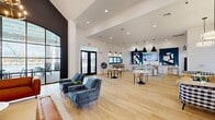 This expansive community clubhouse in Athens, Alabama, delivers a sophisticated blend of modern farmhouse and contemporary design across a generously sized single floor. The grand entrance immediately impresses with soaring vaulted ceilings, a dramatic black accent wall, a large arched steel-frame window flooding the space with natural light, and an elegant multi-ring chandelier overhead. Light-toned hardwood flooring flows seamlessly throughout, unifying each distinct zone.

The open lounge area features rich cognac leather sofas, a wall-mounted flat-screen TV set against a bold teal accent wall, and curated abstract artwork. Adjacent to this is a comfortable seating nook with plush blue velvet armchairs and access to a covered outdoor patio. A well-appointed kitchen and bar area showcases granite countertops, white shaker cabinetry with brass hardware, a stainless steel French door refrigerator, a coffee station, and a patterned tile floor. Pendant lighting and a window bar counter with upholstered stools enhance the casual yet polished atmosphere.

The dining area offers bistro-style tables alongside gingham-upholstered banquettes beneath pendant lights and shiplap-accented wainscoting. A professional reception desk with a dedicated workspace and private hallway connects additional service rooms. Throughout, the design balances openness, warmth, and purposeful functionality.