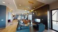 This thoughtfully designed single-floor amenity space offers a sophisticated blend of modern and industrial elements across several distinct yet open zones. Wide-plank hardwood flooring flows throughout, unifying the layout while warm wood cabinetry, exposed brick accents, and dark paneled walls create rich visual contrast.

The main lounge area is anchored by a sleek linear fireplace set into a floor-to-ceiling dark panel wall, surrounded by teal sofas and cognac leather chairs beneath striking gold-and-globe pendant chandeliers. Natural light pours in through large black-framed windows and glass doors that open to an outdoor terrace with panoramic city views.

A generous kitchen and entertaining area features a long white-quartz island, wood cabinetry with stainless steel appliances, pendant lighting, and bold blue tile accents. Framed artwork and a mounted flat-screen television complete the space.

The game room zone includes a full billiards table, a cozy built-in upholstered nook framed in warm wood, and bar-height seating along an exposed brick wall. A separate corner area with floor-to-ceiling windows provides an intimate high-top workspace with sweeping outdoor views. Together, these zones create a versatile, design-forward communal environment.