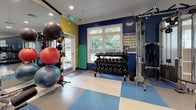 Located at 4937 Valley Crest Dr in Midlothian, VA, this well-equipped fitness center offers a comprehensive workout environment across a single floor. The space is defined by bold navy blue accent walls, vibrant checkered flooring in blue and white, and large white-framed windows that invite abundant natural light and views of the surrounding greenery.

The cardio zone features a row of Life Fitness treadmills, ellipticals, and stair climbers, each equipped with individual display screens for entertainment and workout tracking. Overhead, recessed lighting and ceiling fans ensure the space remains comfortable during intense sessions. Wall-to-wall mirrors amplify the sense of space and allow for form monitoring throughout workouts.

The strength training area is stocked with a full array of Life Fitness selectorized machines, catering to a wide range of muscle groups. A dedicated free weights section includes a complete rack of dumbbells and an assortment of stability and medicine balls, supported by a functional trainer for versatile exercise options. Wall-mounted flat-screen televisions are positioned throughout for entertainment.

Multiple glass-paned doors provide direct outdoor access, enhancing ventilation and connectivity to the exterior. The overall ambiance is energetic yet functional, with a clean, organized layout designed to support a full range of fitness activities.