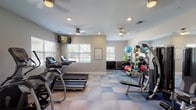 This fitness center is a thoughtfully designed workout facility that offers a comprehensive suite of equipment for a full-body exercise experience. The space features a distinctive checkered floor with blue and neutral tones, providing a clean and energizing aesthetic throughout.

The cardio zone is well-appointed with multiple treadmills and elliptical machines, all positioned to take advantage of the natural light streaming in through large windows fitted with white blinds. Wall-mounted flat-screen televisions are conveniently positioned throughout the room, allowing users to stay entertained during their workouts.

The strength training area includes a multi-station cable machine, a dumbbell rack, and a dedicated free weights section. A large wall mirror amplifies the sense of space and allows users to monitor their form. A well-stocked accessories station holds an assortment of stability balls, resistance bands, and exercise mats, catering to functional and flexibility training.

Ceiling fans and recessed lighting ensure the space remains comfortable and well-lit. The fitness center is accessed through elegant French doors with glass panels, connecting to an adjacent area. Neutral gray walls and white trim contribute to the room's clean, modern appearance, creating an inviting environment conducive to focused, effective workouts.