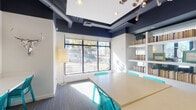 Located at 2501 N Lamar Blvd in Austin, Texas, this single-floor amenity space is designed to support productivity and community engagement with a lively, modern aesthetic. The interior is characterized by a bold navy blue ceiling with exposed ductwork and recessed lighting, contrasting sharply against crisp white walls to create a contemporary industrial atmosphere.

The study lounge serves as a central gathering area, featuring a large communal table surrounded by translucent teal wire-frame chairs. Built-in white shelving units stocked with books line the walls, adding a library-like warmth to the space. Large windows allow generous natural light to flood in, offering views of the surrounding streetscape and mature trees along N Lamar Blvd. A floor lamp adds soft ambient lighting to complement the natural light.

Adjacent to the lounge is a dedicated business center, equipped with desktop computers—including both PC and Mac workstations—arranged along a continuous white built-in desk. A printer is also available for resident use. The entire wall above the workstations serves as a large whiteboard surface, decorated with colorful hand-lettered messaging, announcements, and event information, lending an energetic, campus-style character to the space. Glass partitions connect the areas, maintaining an open and transparent flow throughout.
