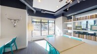 Located at 2501 N Lamar Blvd in Austin, Texas, this single-floor amenity space is designed to support productivity and community engagement with a lively, modern aesthetic. The interior is characterized by a bold navy blue ceiling with exposed ductwork and recessed lighting, contrasting sharply against crisp white walls to create a contemporary industrial atmosphere.

The study lounge serves as a central gathering area, featuring a large communal table surrounded by translucent teal wire-frame chairs. Built-in white shelving units stocked with books line the walls, adding a library-like warmth to the space. Large windows allow generous natural light to flood in, offering views of the surrounding streetscape and mature trees along N Lamar Blvd. A floor lamp adds soft ambient lighting to complement the natural light.

Adjacent to the lounge is a dedicated business center, equipped with desktop computers—including both PC and Mac workstations—arranged along a continuous white built-in desk. A printer is also available for resident use. The entire wall above the workstations serves as a large whiteboard surface, decorated with colorful hand-lettered messaging, announcements, and event information, lending an energetic, campus-style character to the space. Glass partitions connect the areas, maintaining an open and transparent flow throughout.