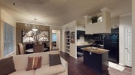 This well-appointed single-floor residence showcases a thoughtfully designed open-concept layout that seamlessly connects the living, dining, and kitchen areas into one cohesive and functional living space. Rich, dark hardwood flooring flows continuously throughout, anchoring the warm, neutral color palette of cream and mocha tones that define the interior's refined aesthetic.

The kitchen is a standout feature, equipped with sleek dark cabinetry, granite countertops, and black appliances including a refrigerator, microwave, and range. A mosaic tile backsplash adds a decorative focal point, while a peninsula counter provides additional prep space and casual seating. Crown molding trims the upper cabinetry, lending an elevated, polished finish.

Adjacent to the kitchen, a built-in shelving unit offers practical display and storage space, serving as a subtle architectural divider between the kitchen and living zones. The living area is bright and welcoming, centered around a plush cream sofa accented with decorative pillows.

The dining area is distinguished by a stylish pendant light and wall art, creating an intimate yet open atmosphere. A hallway beyond provides access to additional private rooms. Throughout the space, crown molding, consistent finishes, and cohesive design choices contribute to a refined and comfortable ambiance.