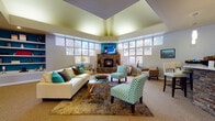 This single-floor communal lounge is designed to impress, combining architectural interest with a warm, inviting atmosphere. The space is anchored by a striking corner fireplace clad in stacked stone, topped with a mounted flat-screen television and set against a bold teal accent wall. Surrounding the fireplace, expansive floor-to-ceiling windows wrap around the room, flooding the interior with abundant natural light and offering views of the surrounding exterior.

The ceiling is a notable architectural feature, rising to a dramatic tray design with tiered levels that add depth and visual height to the room. Warm recessed and ambient lighting complements the natural light throughout the day. The main seating area features a cream sofa accented with teal and earth-toned throw pillows, paired with chevron-patterned accent chairs and a glass-topped coffee table set over a layered area rug.

Toward one side of the space, a built-in bookshelf with a teal backing provides both display and storage. A separate bar area with a stacked-stone base, light countertop, pendant lighting, and white leather bar stools offers a functional and stylish gathering spot. A chandelier-lit corridor connects the lounge to additional areas, rounding out this well-appointed space.