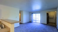 This single-floor space offers a straightforward, functional layout designed for comfortable everyday living. The main living area is open and airy, featuring plush wall-to-wall carpeting that adds warmth underfoot. A large sliding glass door serves as the focal point of the living space, flooding the room with natural light and providing direct access to an outdoor balcony. Sheer curtains frame the door, softening the natural light that pours in throughout the day. A baseboard heating unit runs along the exterior wall beneath the sliding door, ensuring efficient climate control throughout the space. The living area flows naturally into a kitchen zone, partially separated by a raised counter with a laminate surface — ideal as a breakfast bar or casual dining space. Crisp white walls throughout contribute to a clean, neutral aesthetic that is easy to personalize. A hallway visible from the living area leads to additional rooms, suggesting a well-organized floor plan. The smooth, flat ceilings maintain a consistent and uncluttered look throughout the space. Overall, this unit offers a practical and welcoming environment with key features including balcony access, natural light, and an open-concept connection between the kitchen and living areas.