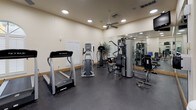 This fitness center offers a comprehensive workout environment designed to accommodate a wide range of exercise routines. The space is finished with durable rubber flooring throughout, providing a safe and functional surface for all types of training activities. Large wall-mounted mirrors span multiple sections of the facility, visually expanding the space and allowing users to monitor their form during workouts.

The cardio zone features a row of True-brand treadmills and elliptical machines, positioned near arched windows fitted with plantation shutters that invite natural light into the space. A dedicated free weights area includes a full rack of dumbbells in varying sizes, accompanied by a weight bench, a stability ball, and balance training accessories. The strength training section is equipped with a multi-station cable machine, offering versatility for a broad spectrum of resistance exercises.

Ceiling fans complement the recessed lighting throughout, maintaining comfortable airflow during workouts. Wall-mounted flat-screen televisions are positioned for visibility across the floor. The facility is accessed through glass-panel double doors and maintains a clean, neutral aesthetic with warm beige walls and wood-toned baseboards. Decorative potted plants add a touch of warmth to the otherwise functional environment, creating an inviting atmosphere for daily fitness routines.