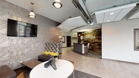 The Commons is a thoughtfully designed single-floor space that blends modern aesthetics with an inviting, communal atmosphere. At its heart is a reception area anchored by a sleek, dark reception desk set against a striking reclaimed wood accent wall bearing "The Commons" signage, immediately establishing a warm yet professional identity upon entry.

The lounge area is defined by a textured, concrete-style accent wall that serves as a sophisticated backdrop for a wall-mounted flat-screen display. A series of pendant lights with polished metal fixtures hang at varying heights, providing layered illumination throughout the space. Seating is arranged to encourage both casual interaction and individual use, with low-profile upholstered sofas and ottomans in neutral tones complemented by patterned accent pieces in yellow, gray, and white. Low wooden coffee tables and mixed seating, including bentwood chairs and round white tables, add variety and functionality.

Exposed ductwork runs along the ceiling, contributing to the space's industrial character, while patterned carpet tiles ground the lounge zone. Light tile flooring near the reception area creates a clear visual transition between zones. The overall ambiance is modern, open, and professionally curated.