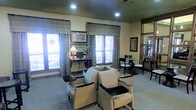 This single-floor space is thoughtfully designed to serve as a multifunctional common area, combining a lounge, workstation, and dining zone within one cohesive environment. The room is finished with neutral-toned walls and carpeted flooring, contributing to a warm and comfortable ambiance throughout.

At the center of the space, two cushioned armchairs with dark wooden armrests are arranged to encourage conversation and relaxation. Adjacent to the seating area, a dedicated computer workstation features a desk, monitor, and a table lamp, providing a functional area for residents or visitors to work or browse. A framed artwork piece hangs above the desk, adding a decorative touch.

Multiple windows fitted with woven bamboo-style curtains and horizontal blinds line the walls, allowing generous natural light to fill the room while maintaining privacy. The dining area, positioned toward the back, includes a glass-topped table surrounded by dark wooden chairs, ideal for small group gatherings.

Recessed ceiling lighting supplements the natural light, ensuring the space remains well-illuminated. A mirrored accent and additional framed artwork on the walls enhance the room's sense of depth and visual interest, completing its welcoming, community-oriented character.