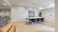 Located at 2132 Wilkinson St in Kelowna, BC, this thoughtfully designed community lounge offers a versatile, single-floor layout that balances comfort and functionality. The space is finished with light hardwood-style flooring throughout, crisp white walls, and recessed lighting that creates a clean, welcoming ambiance.

The living area serves as the social heart of the space, furnished with contemporary seating including a cognac leather armchair and a sleek black leather accent chair arranged around a wall-mounted flat-screen television. A sideboard and framed black-and-white photography add curated character to the room, while windows allow natural light to filter in.

Adjacent to the lounge is a well-equipped kitchen featuring two-tone cabinetry — warm wood-grain lower cabinets paired with white upper cabinets — complemented by white countertops and stainless steel appliances including a refrigerator, dishwasher, and range. The galley-style layout is efficient and professionally appointed.

A separate meeting and dining area rounds out the space, centered around a large conference-style table with seating for up to eight. Pendant lighting hangs above the table, and a mounted display screen and a wall-mounted climate control unit add practical functionality. A potted fiddle-leaf fig plant brings a touch of greenery to the room. The space accommodates a maximum occupancy of 30 people.