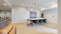 Located at 2132 Wilkinson St in Kelowna, BC, this thoughtfully designed community lounge offers a versatile, single-floor layout that balances comfort and functionality. The space is finished with light hardwood-style flooring throughout, crisp white walls, and recessed lighting that creates a clean, welcoming ambiance.

The living area serves as the social heart of the space, furnished with contemporary seating including a cognac leather armchair and a sleek black leather accent chair arranged around a wall-mounted flat-screen television. A sideboard and framed black-and-white photography add curated character to the room, while windows allow natural light to filter in.

Adjacent to the lounge is a well-equipped kitchen featuring two-tone cabinetry — warm wood-grain lower cabinets paired with white upper cabinets — complemented by white countertops and stainless steel appliances including a refrigerator, dishwasher, and range. The galley-style layout is efficient and professionally appointed.

A separate meeting and dining area rounds out the space, centered around a large conference-style table with seating for up to eight. Pendant lighting hangs above the table, and a mounted display screen and a wall-mounted climate control unit add practical functionality. A potted fiddle-leaf fig plant brings a touch of greenery to the room. The space accommodates a maximum occupancy of 30 people.