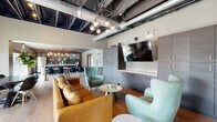 Located at 20 S Naper Blvd in Naperville, Illinois, this single-floor space blends industrial design with modern comfort to create a dynamic and inviting environment. The open layout features exposed ductwork and an open ceiling with bold pendant lighting, lending a distinctive industrial-chic character throughout.

The main lounge area is anchored by a rich tan leather sectional sofa, complemented by teal accent chairs and round metallic side tables, all set atop warm hardwood flooring. A large wall-mounted flat-screen TV is flanked by a full wall of sleek grey cabinetry, offering ample storage. A glass-partitioned room adjacent to the lounge provides a more private meeting or conferencing option.

The kitchen is a standout feature, boasting dark cabinetry with warm wood accents, a textured dark tile backsplash, exposed wooden ceiling beams, and a generous white quartz island with bar-height seating. Glass pendant lights in brushed brass add a refined touch, while stainless steel appliances, including a full-size refrigerator, ensure full functionality.

A dedicated dining and work zone features a solid wood communal table with modern black chairs, alongside a standing-height workstation with power access. The space transitions smoothly between social, collaborative, and productive uses, making it a well-rounded, thoughtfully designed hub.
