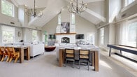 Located at 56 Republic Dr in Bloomfield, CT, this single-floor space makes a striking first impression with its dramatic vaulted ceilings adorned with exposed beam detailing and dual wrought-iron chandeliers. The expansive main gathering area centers around a floor-to-ceiling whitewashed stone fireplace with a rustic wood mantel, flanked by generously sized sofas and complemented by a solid wood dining table with woven rattan chairs. Soft neutral tones, plush carpet, and abundant natural light from multiple windows create a warm, inviting atmosphere throughout. An adjacent lounge area transitions to natural stone tile flooring and features a built-in countertop workspace along a wall of windows, wall-mounted sconce lighting, and a flat-screen TV—ideal for both relaxation and productivity. The well-equipped kitchen includes rich dark wood cabinetry, white quartz countertops, stainless steel appliances, subway tile backsplash, and teal open shelving that adds a bold accent. A dedicated utility or prep room with coordinating teal shelving and additional cabinetry provides practical storage. Throughout, the design blends contemporary farmhouse aesthetics with functional, thoughtfully curated details, resulting in a cohesive and welcoming environment.